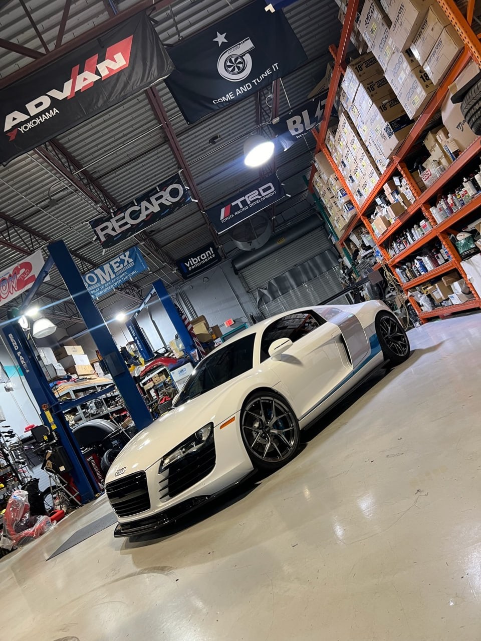 White Audi R8 with a blue livery stripe and dark aftermarket wheels on the Velocity Factor shop floor under Advan, Recaro and TRD banners