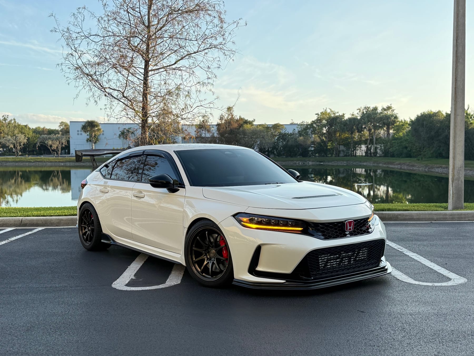 White Honda Civic Type R FL5 on bronze wheels with red Brembo calipers, a front lip and an aftermarket rear wing, parked beside a lake at sunset