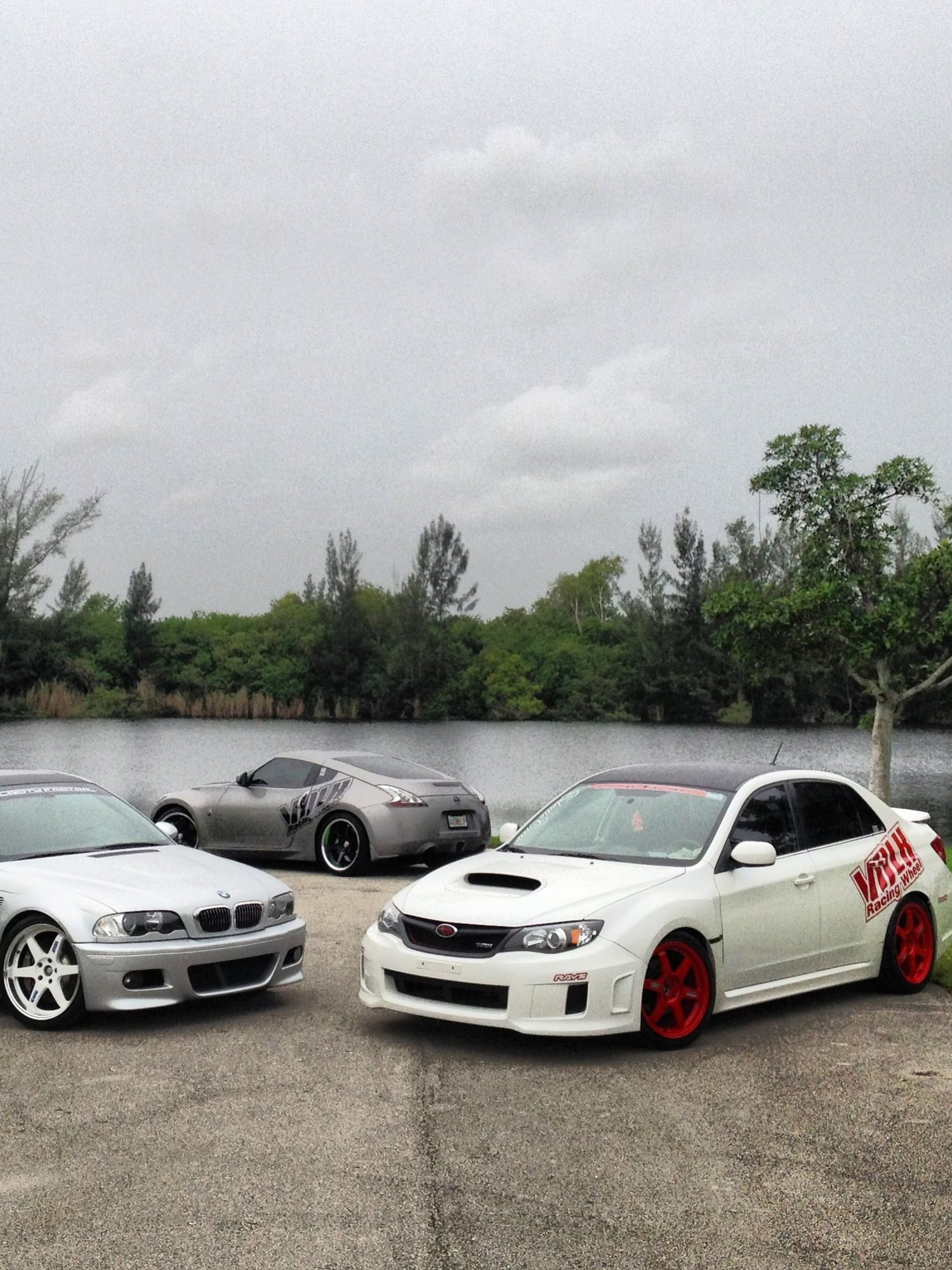White Subaru WRX sedan on red Volk Racing TE37 wheels parked lakeside with a BMW M3 and Nissan 370Z